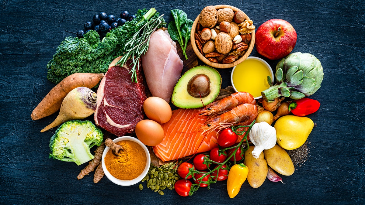 Overhead view of a large group of healthy foods used in a Paleo diet like beef, chicken meat, salmon, sardines, shrimps, root vegetables, greens, vegetables, fruits, extra virgin olive oil, nuts and seeds.