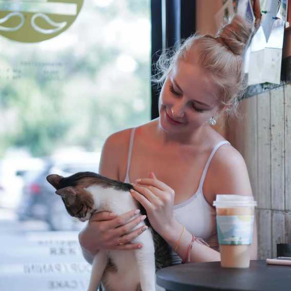 A woman with blonde hair in a bun wearing a white tank top sits by a window holding a tabby cat. A coffee cup is on the table nearby.