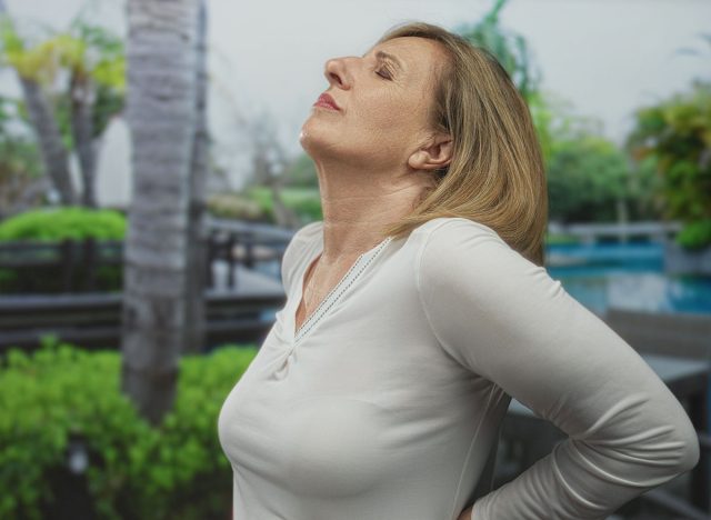 Middle age woman holds lower back and leans backward in green park by poolside chairs on sunny morning; discomfort.