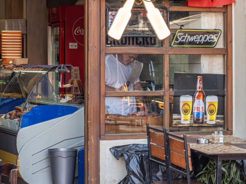 A chef chops food with a large knife behind a window in a restaurant. Bottles of Pils beer and large cups are displayed on the counter outside. A Coca-Cola fridge and a blue ice cream freezer are also visible.