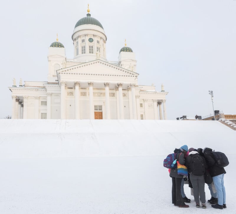 A group of five people with backpacks huddle together in front of the snow-covered Helsinki Cathedral, a large white building with green domes and golden accents.