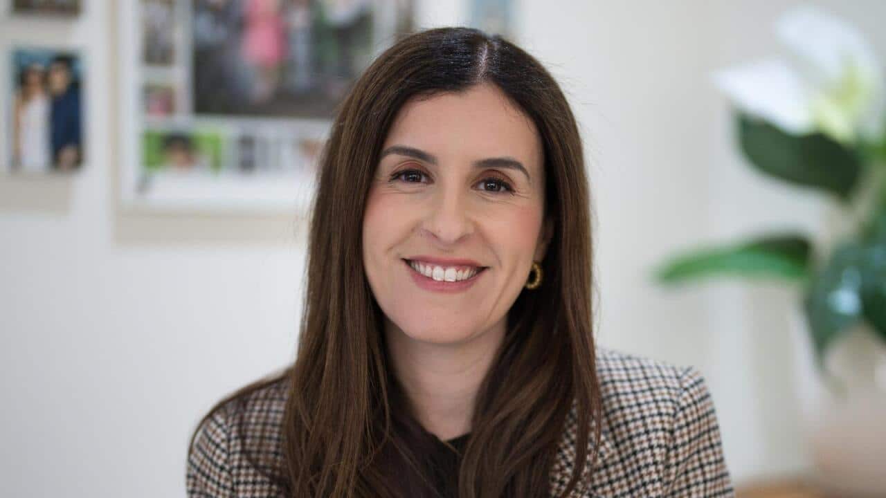 A woman with long black hair, wearing a checkered blazer, is smiling.