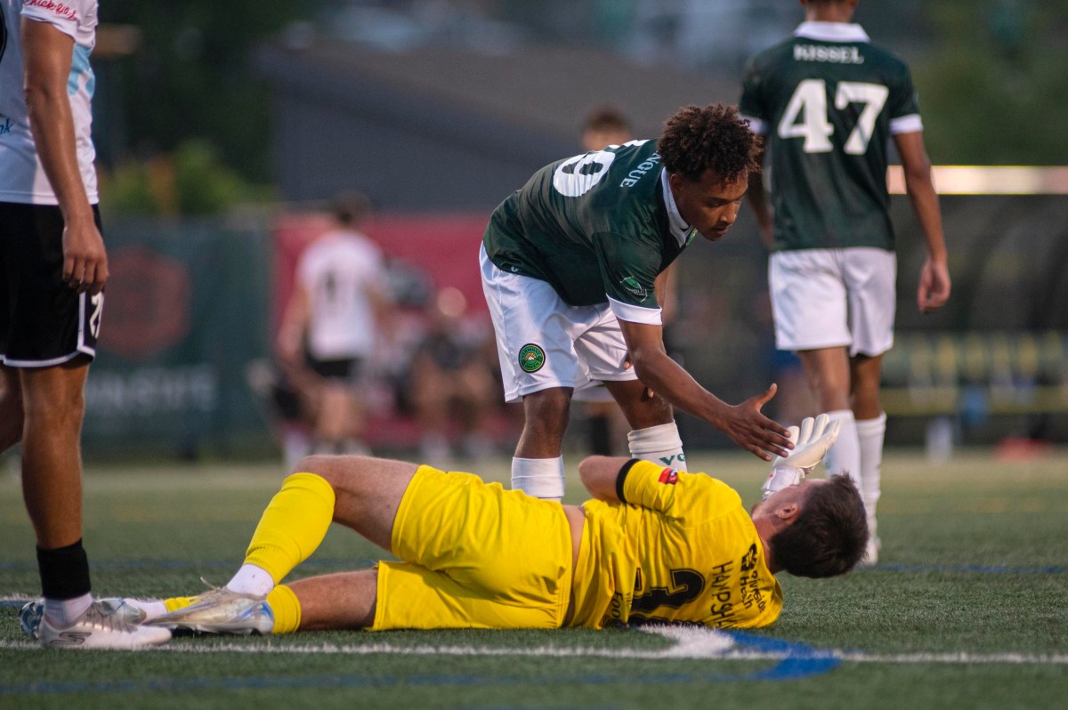 A soccer player in a green jersey helps a goalkeeper in a yellow uniform who is lying on the field during a match.