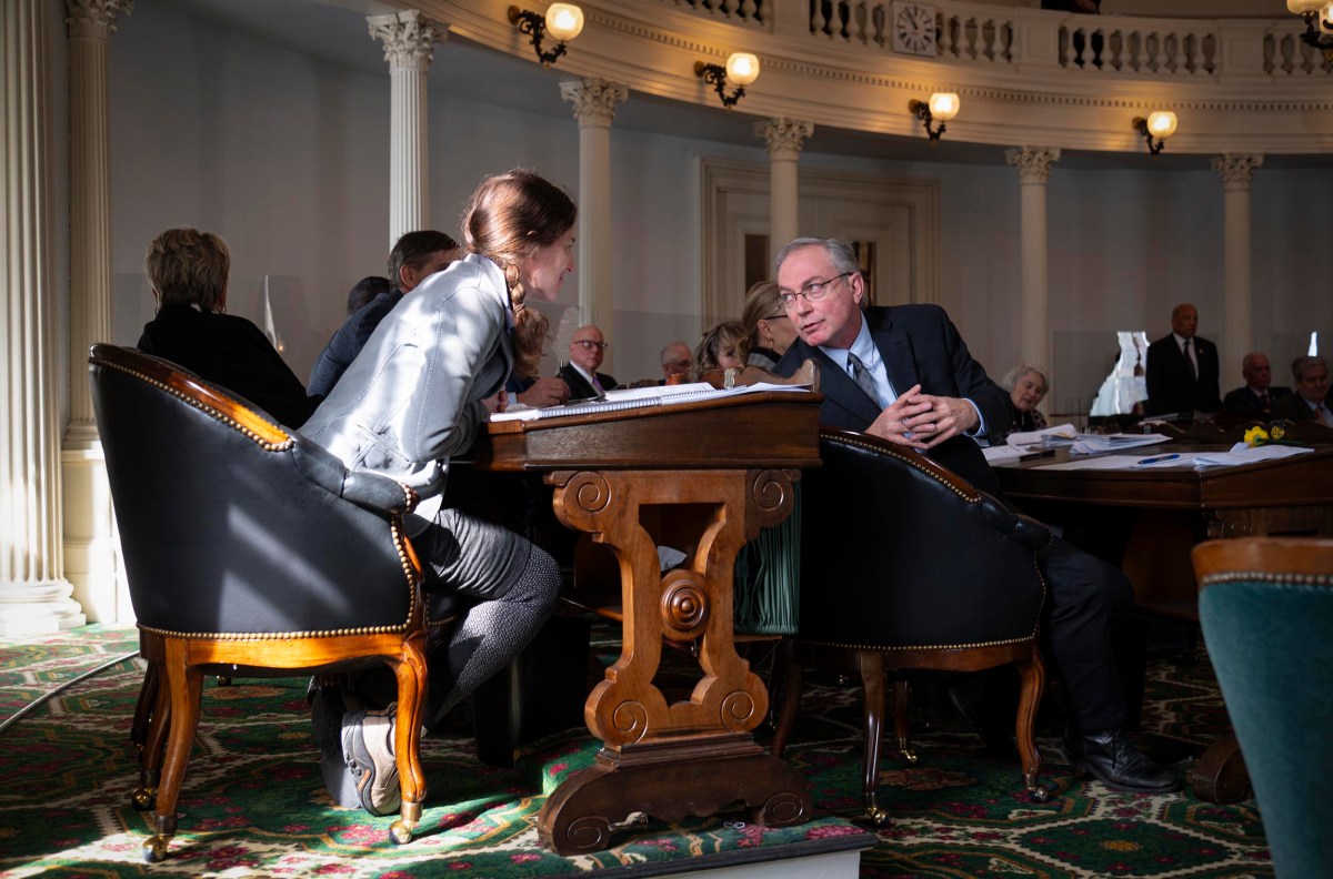 A man and a woman sit at a wooden table in a large, ornate room having a discussion. They are surrounded by other seated individuals.