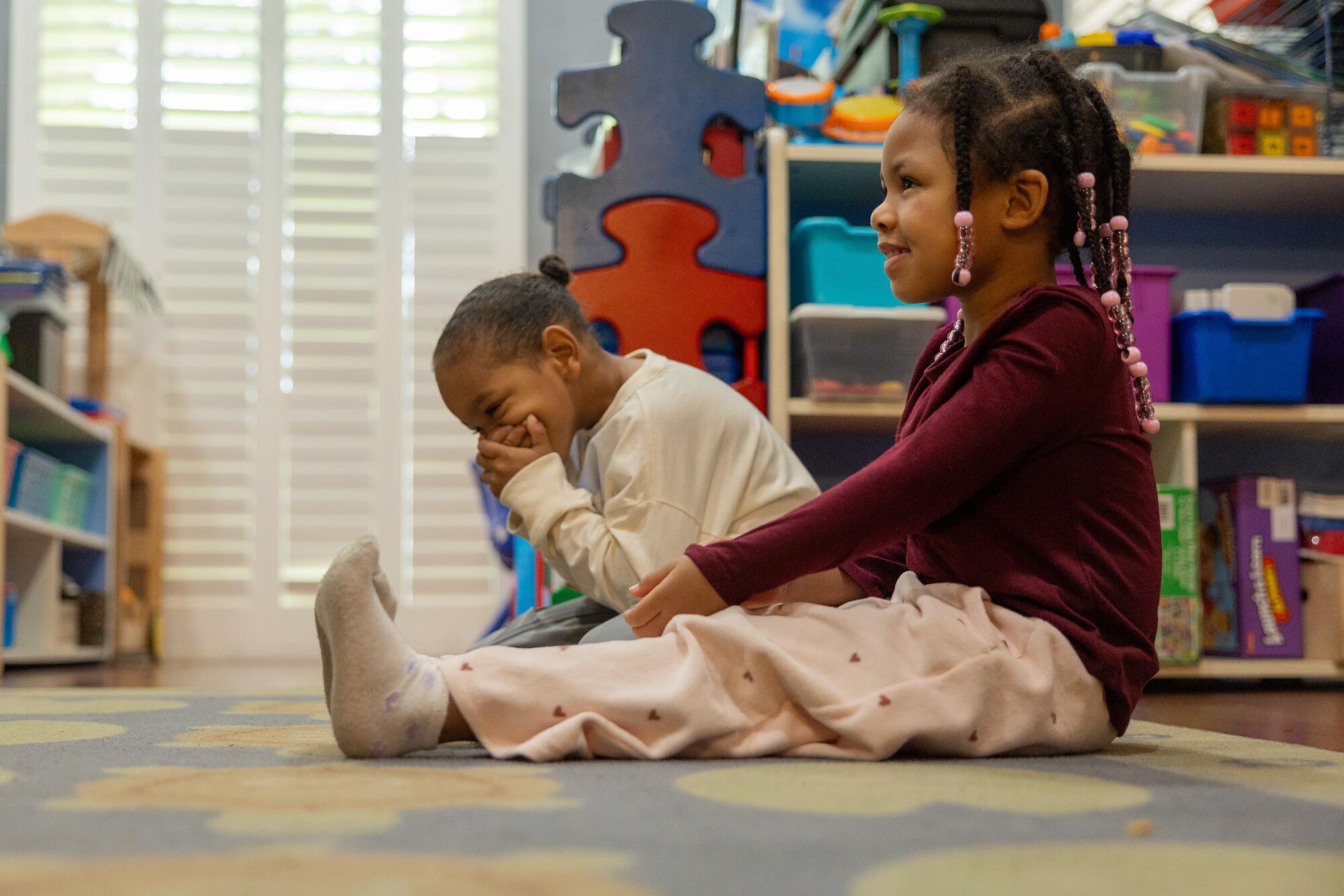 Two children sit on the rug listening as someone speaks. One covers his face with his hands, holding in laughter.