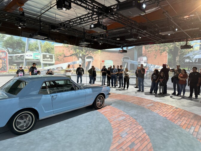 Several people gather around an old blue Mustang inside an art exhibition.