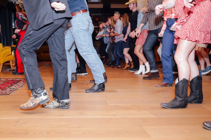 Two lines of men and women, most wearing cowboy boots, are dancing on a wooden floor