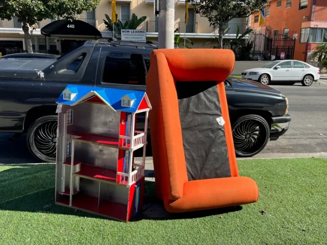 An orange couch leans on a large toy house on a patch of turf grass near a street curb next to a black pick up truck.