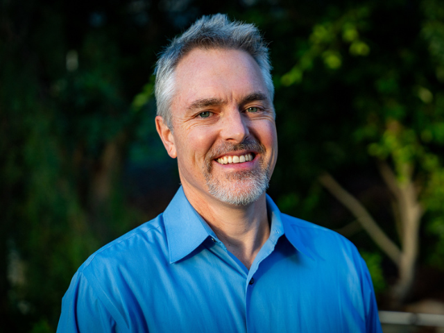 A headshot of a man in a blue shirt in front of a green background.