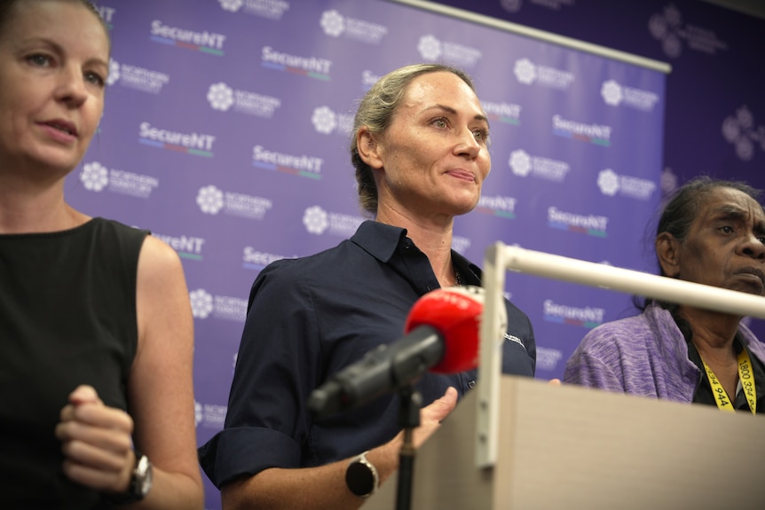 A white woman, blonde hair, navy button up shirt standing at podium, with two women standing either side of her.