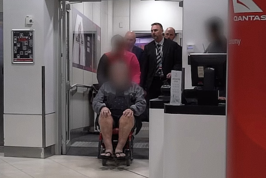 A man with a digitally blurred face is wheeled through the arrivals area of an airport. 