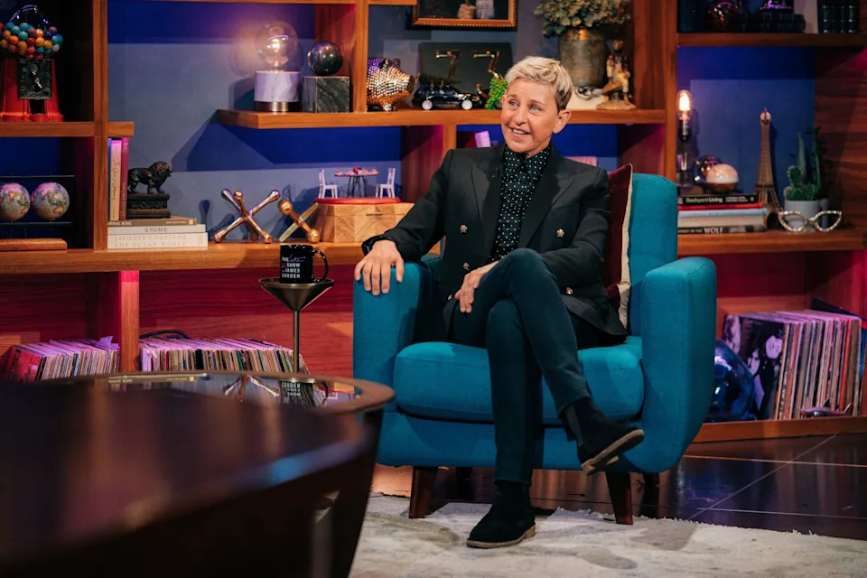 Ellen DeGeneres in tailored suit sits casually in a talk show setting, surrounded by books and decor
