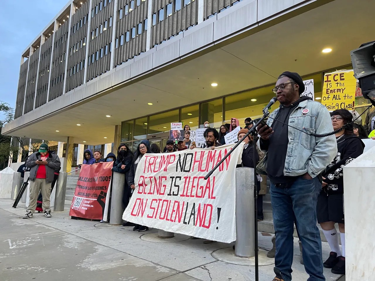 Large group of people standing in front of a large, rectangular building, holding signs and banners. The largest banner says "Trump no human being is illegal on stolen land!" in all caps. In the foreground, a man stands in front of the group on the right side of the image, speaking into a microphone.