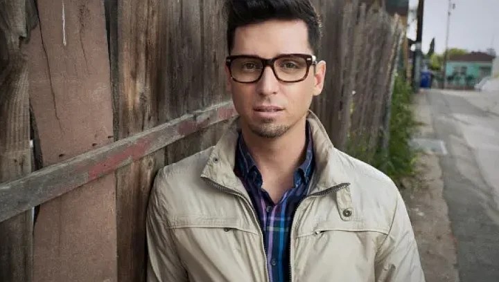 A man with glasses stands next to an old wooden fence