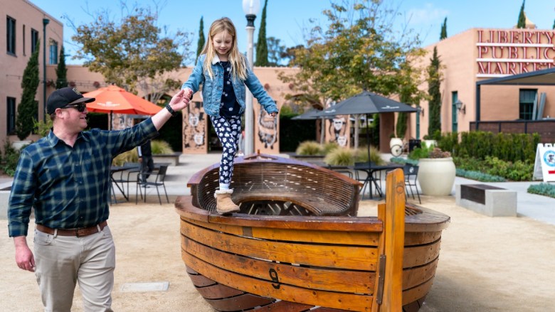 A man helps a girl balance on a wooden boat-shaped structure at Liberty Public Market.
