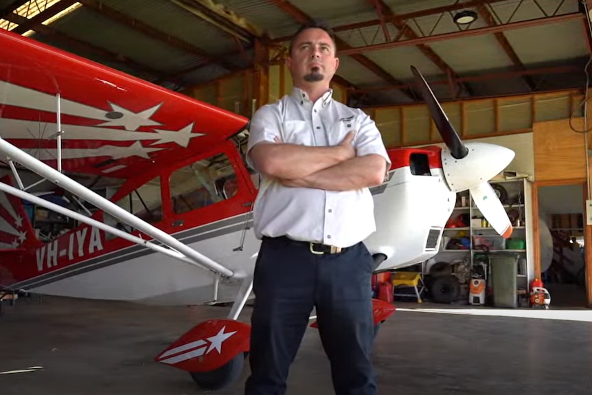 A man in a pilot's shirt stands with his arms crossed in front of a light aircraft in a small hanger