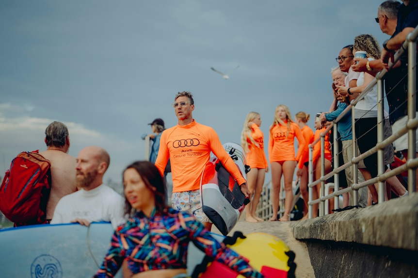 A man with a surfboard goes down steps onto the beach as people leaning against a railing watch the event.