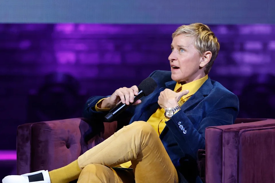 Ellen DeGeneres in stylish suit sitting with a microphone, gesturing while speaking on stage