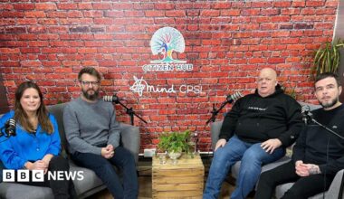 Members of the CPSL podcast team- a woman in a blue blouse, a man in a grey jumper and navy trousers, a man in a black hoodie and blue jeans and a man in a black jumper and black trousers- sitting on grey sofas on either side of the room with microphones in front of them.