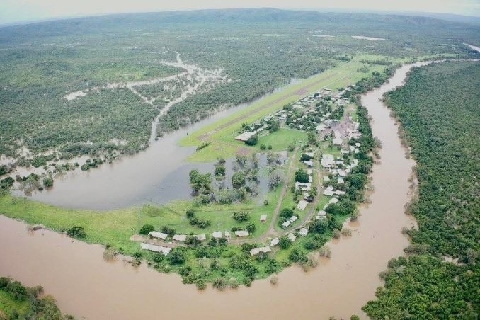An aerial image of a full river, leaking onto green land with homes. Green bushes surrounding the river.