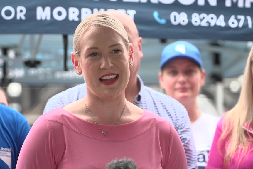 a blonde woman in a pink top speaks at a press conference