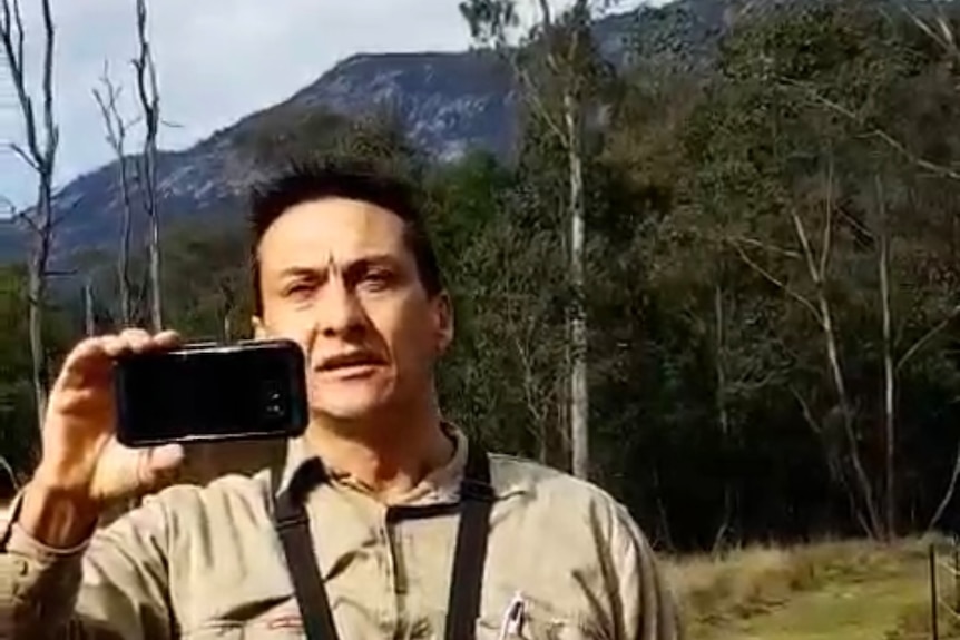 A dark-haired man holds his phone while standing in a tree-fringed paddock in front of a mountain range.