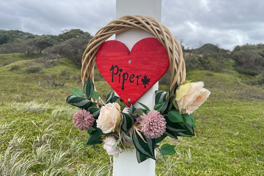 A red wooden heart saying "Piper" hangs from a cross in coastal shrubland.