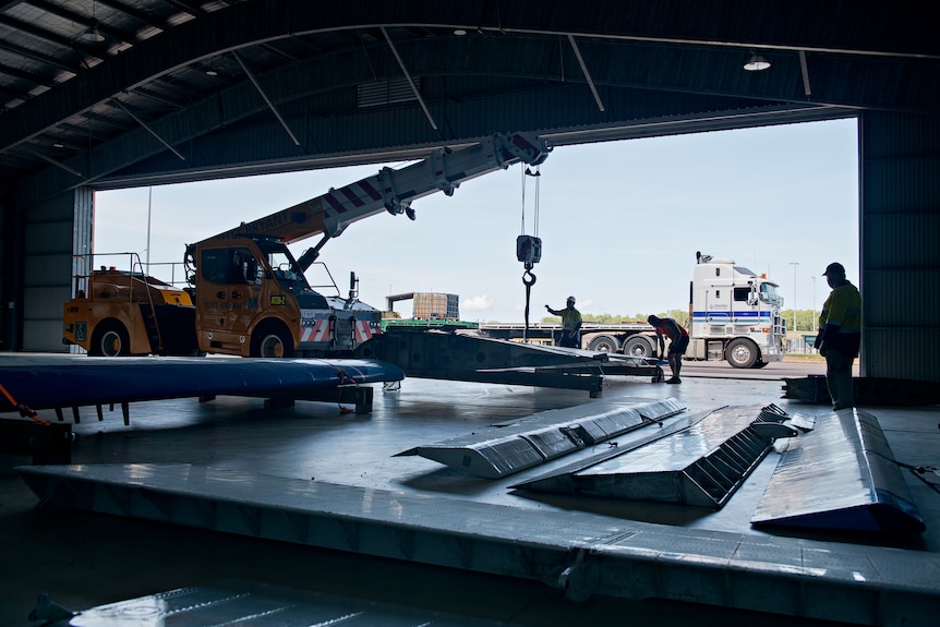 A shadowy image taken at the back of industrial shed / plane hangar, with plane parts being lifted in, workers around.