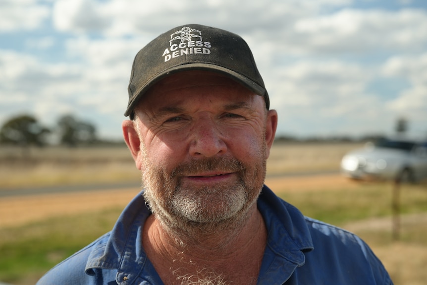 Close up portrait of male farmer looking down barrel of camera with farm in the background, serious expression, wearing cap
