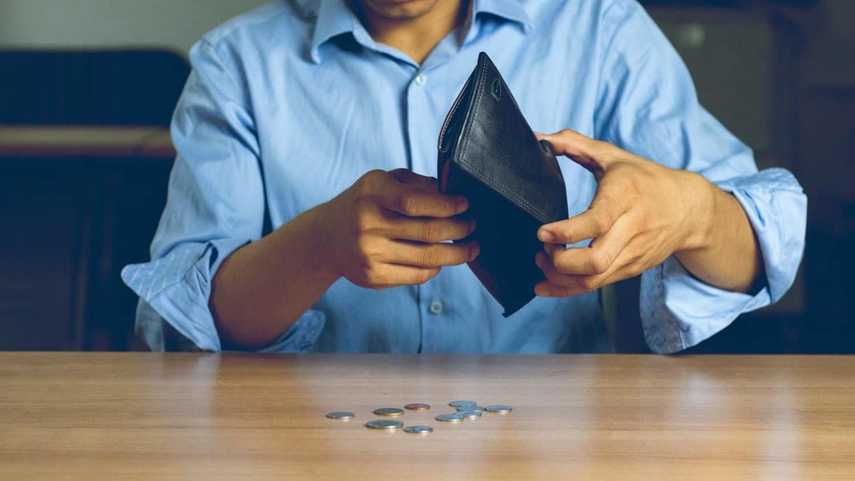 A person, wearing a light blue button-down shirt, holds an open, empty black wallet over a wooden table. A small scattered pile of various silver and copper coins lies on the table in front of the wallet, indicating financial hardship.
