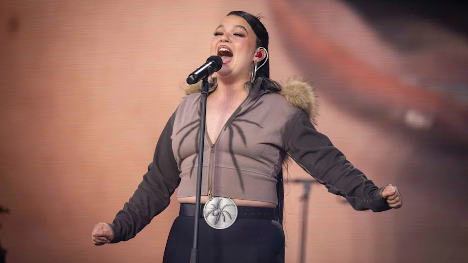 Lola Young is singing into a microphone on stage. She wears a brown zip up jacket with a fur collar hood paired with black trousers.