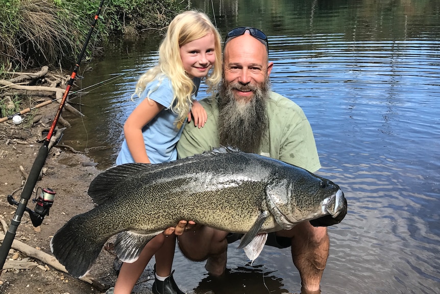 Man with beard and little girl holding a 1m long Murray cod by a river bank.