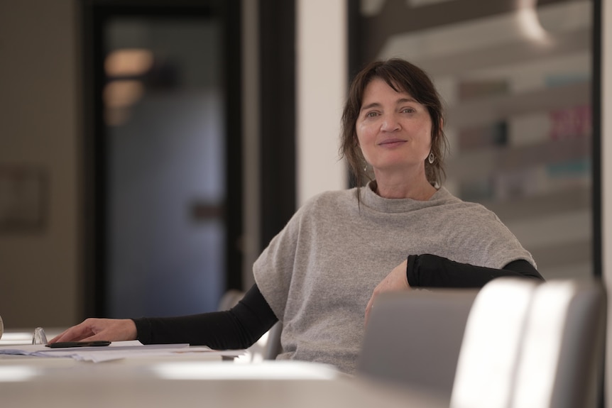 A woman with brown hair sits at a table with her arm rested on the back of the chair, looking at camera.
