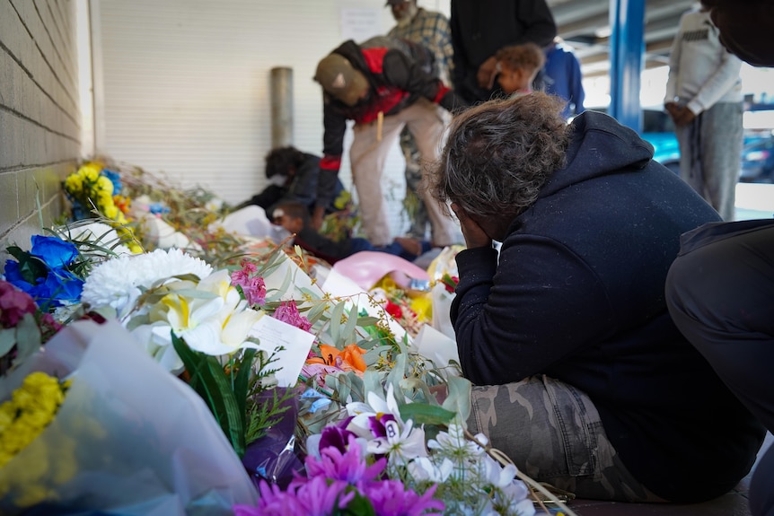 A woman bends over in mourning next to a pile of flowers laid on the ground.