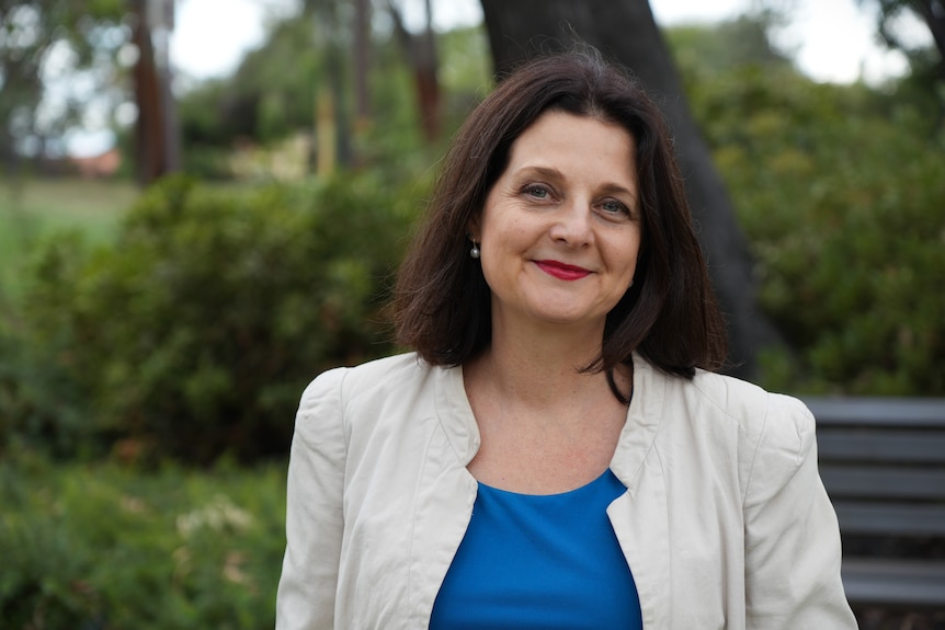 Woman wearing blue shit and white blazer stands in front of tree.