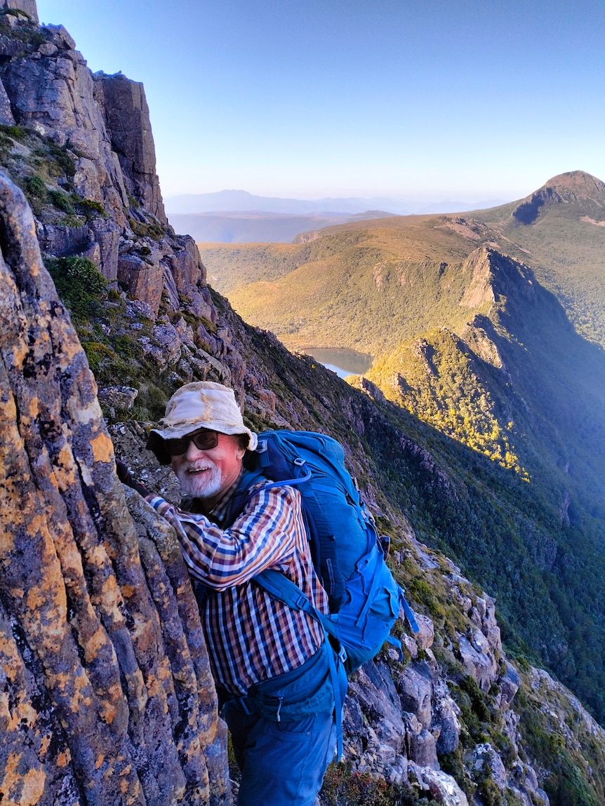 A vertical photo of an older man holding on to the side of a cliff, with a steep drop into a valley below