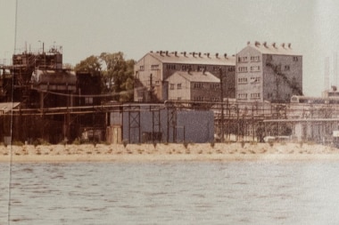 A dull old photograph of three large multi-storey buildings by water in the bay with industrial infrastructure next to it.