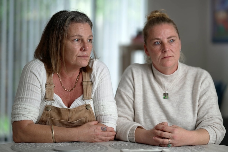 Two women sit at a table, with serious, reflective expressions.