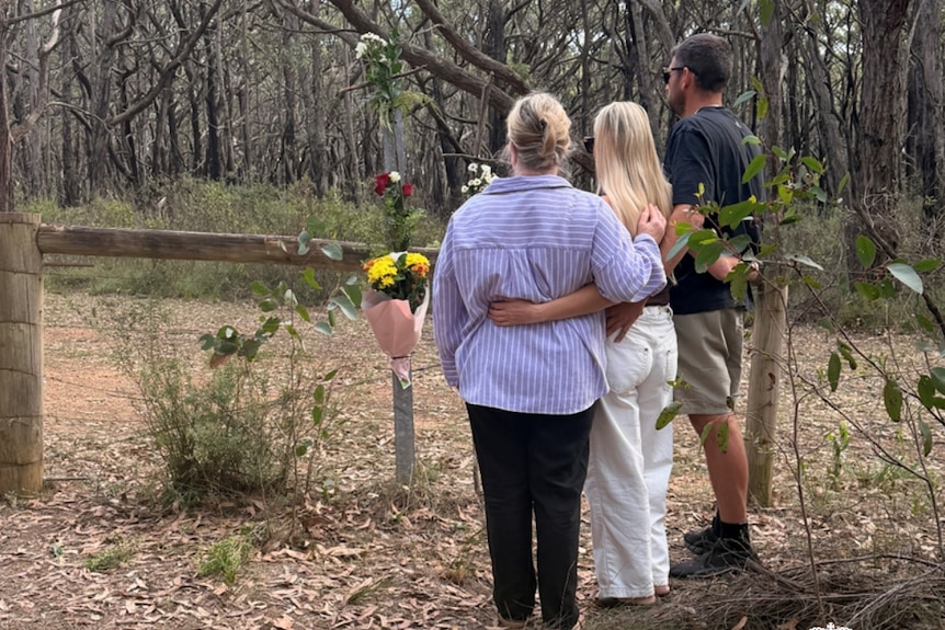Two women and a man stand near a fence with a bouquet of flowers attached to it, with trees behind