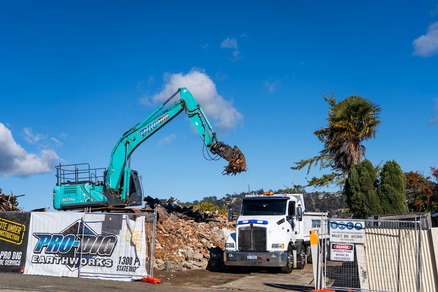 a blue excavator digs at a pile of old timber and brick