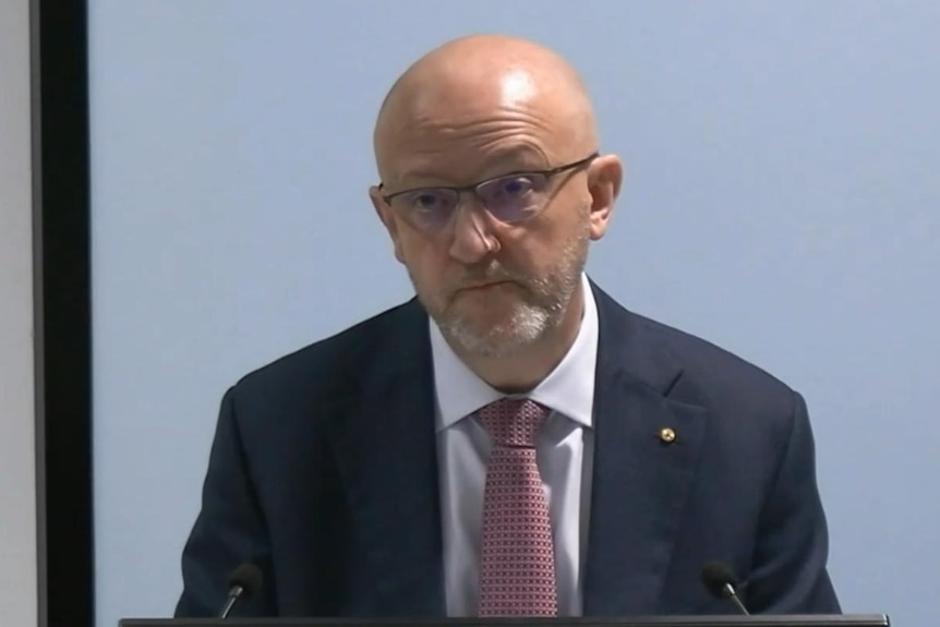 Man in suit and glasses behind microphone, grey background
