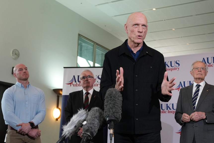 Peter Garrett at a press conference, wears black, three men, two in suits behind him.