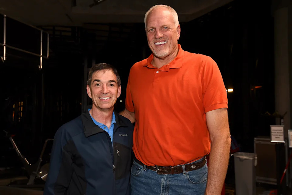 Two men standing together, smiling. One is wearing a casual jacket, and the other is in a polo shirt. They appear to be in a sports setting