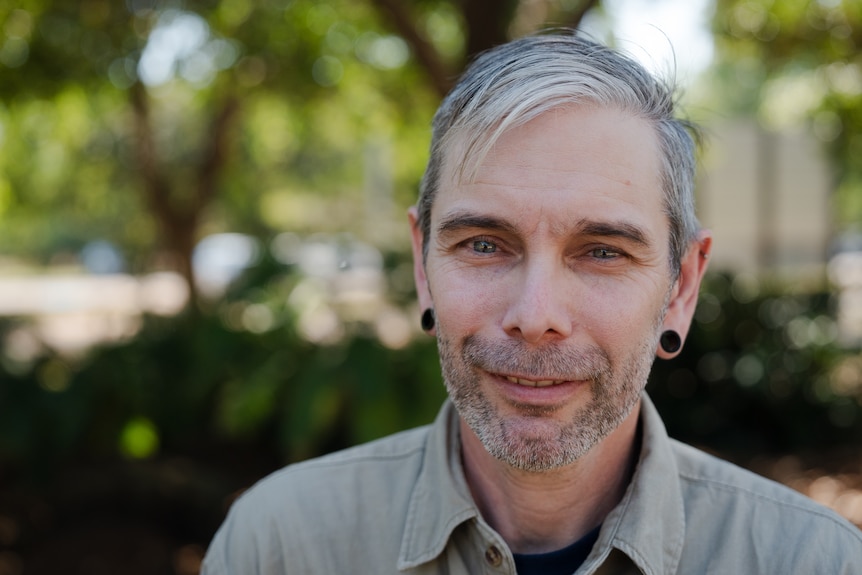 A man with light hair smiling at the camera, green trees can be seen in the background.