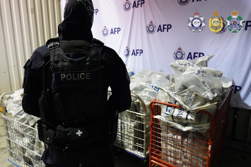Back of a police officer dressed head to toe in black, with orange and white trolleys filled with parcels in bags.