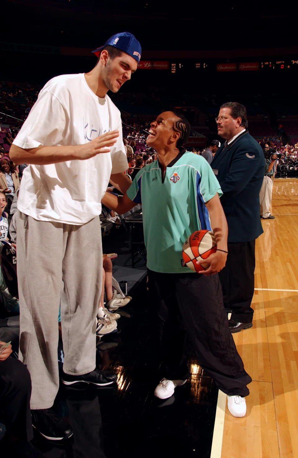 Two basketball players, one much taller, are interacting on a court. The shorter player holds a basketball, wearing a sports polo and pants