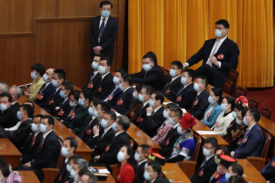People seated in an auditorium wearing face masks, with some individuals standing. The setting suggests a formal assembly or conference