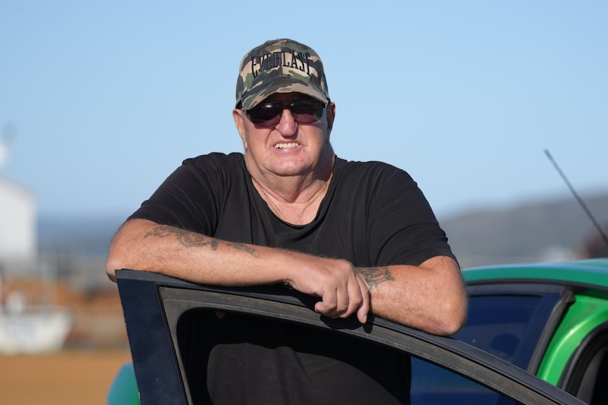 A man wearing sunglasses, a cap and a dark T-shirt leans on a car door.