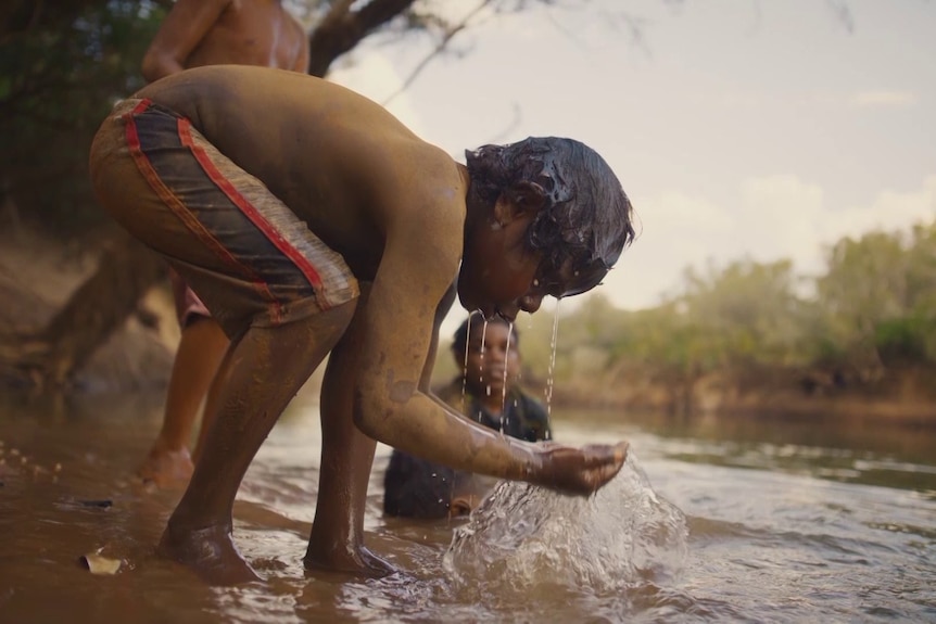 A young boy scoops water from the river to wash his face while others look on