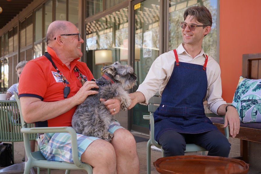 Two men sit outside a cafe smiling at each other. One is holding a dog on his lap, the other wears an apron.